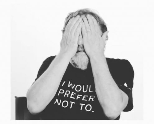 Slavoj Zizek covering his eyes while wearing a t shirt that says "I would prefer not to"
